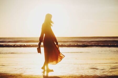 young slim beautiful woman on sunset beach, playful, dancing, running, bohemian outfit, indie style, summer vacation, sunny, having fun, positive mood, romantic, splashing water, silhouette, happyの写真素材