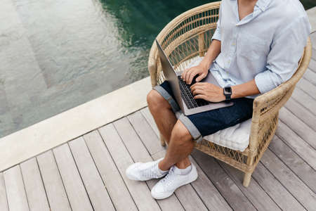 Young man using a laptop computer in a garden with a swimming pool.
