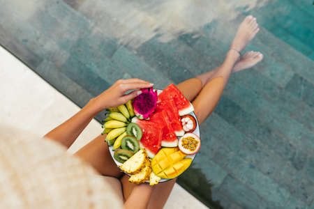 Young unrecognizable woman by the pool with a plate of tropical fruits: watermelon, pineapple,...