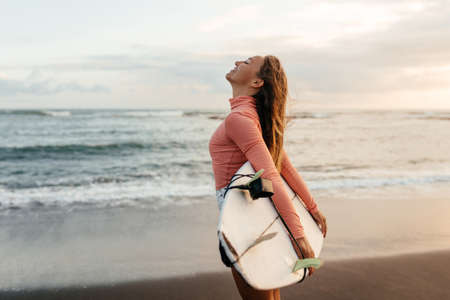 Young attractive surfer woman with white board at sunset on the ocean. Bali Indonesia.