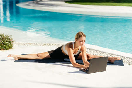 Young fit woman doing yoga stretching exercise outdoor near swimming pool in front of laptop.