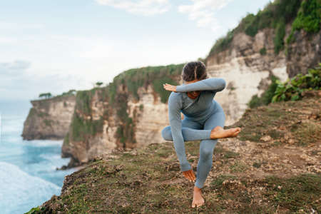 Beautiful woman doing yoga on a cliff, behind an amazing view in the oceanの写真素材
