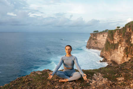 Beautiful woman doing yoga on a cliff, behind an amazing view in the oceanの写真素材