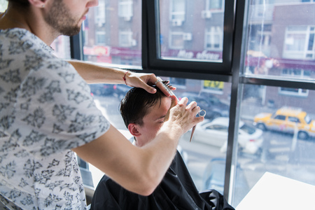 A professional hairstylist with a comb and scissors in his hand styling the wet black and short hair of the man in a hair salonの写真素材