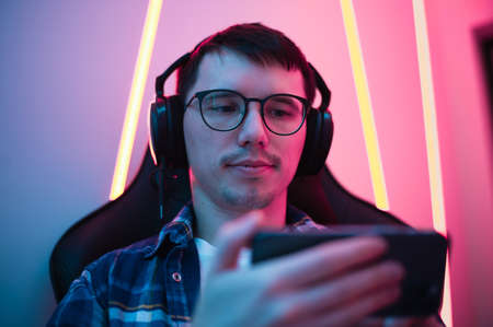 Close up portrait of a boy relaxing playing games on mobile phone and sitting in a gaming chair. Room with colorful neon lightの写真素材