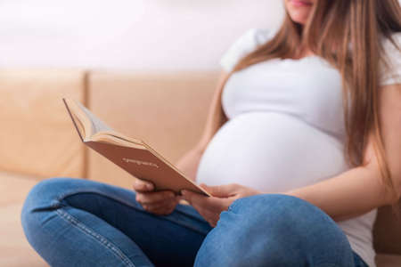 a pregnant girl sits on the couch and reads a book about pregnancyの写真素材