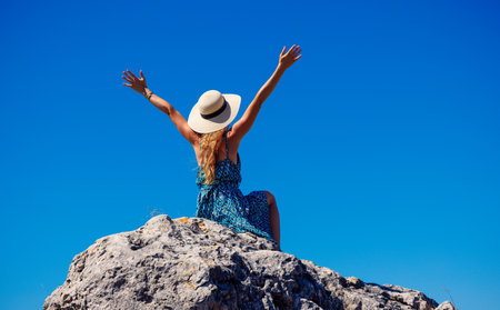 Woman with hat and blue dress sitting on peak on blue sky- vacation, freedom, happiness conceptの写真素材
