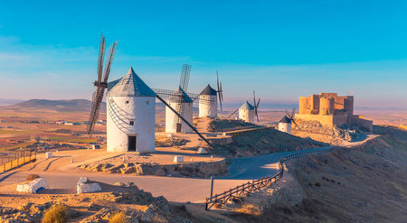 Traditional windmills and a medieval castle on the hill of Consuegra, Spain, illuminated by the golden light of sunset, evoking the legendary landscapes of Don Quixote.の写真素材
