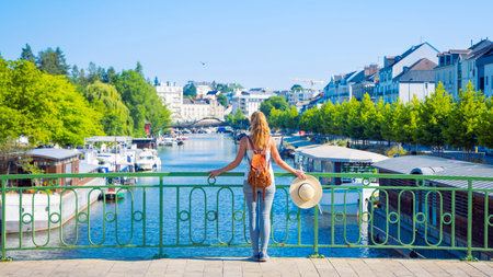 A woman with a backpack and hat stands on a bridge overlooking the river in Nantes, France, admiring the scenic urban waterfront with boats and greenery.の写真素材