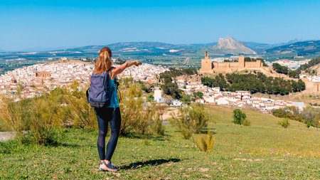 A female traveler with a backpack points towards the historic fortress of Alcazaba in Antequera, MÃ¡laga, Spain, overlooking the whitewashed town and surrounding landscapesの写真素材