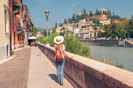 A woman in casual summer attire walks along a scenic riverside promenade in Verona, Italy, admiring the historic architecture and lush hillside.の写真素材