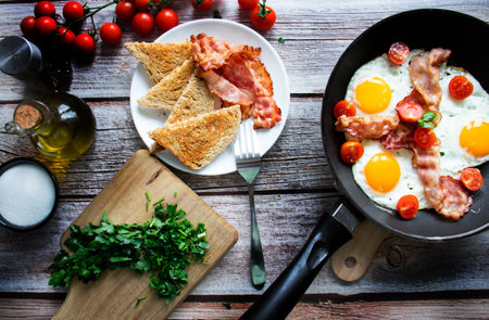 Fried eggs with bacon and tomatoes in a frying pan on a wooden table