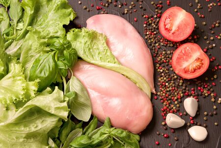 Chicken fillet next to greens spinach, peppercorns, cherry tomatoes and garlic on a black background. Dietary meat. Proper nutritionの写真素材