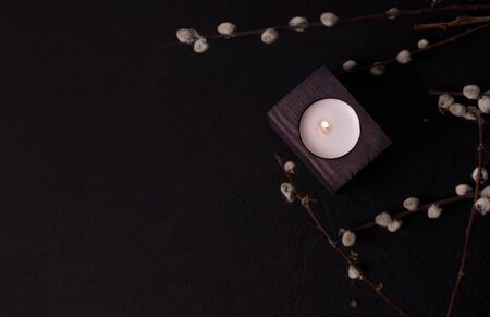 one candle in a wooden candlestick in willow branches on a black background. dark environmental...