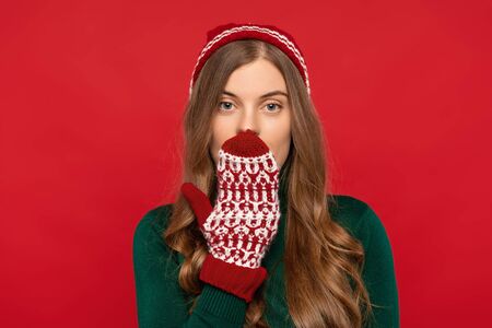 front view of beautiful woman in winter hat and mittens covering mouth with hand isolated on red background. Christmas conceptの写真素材