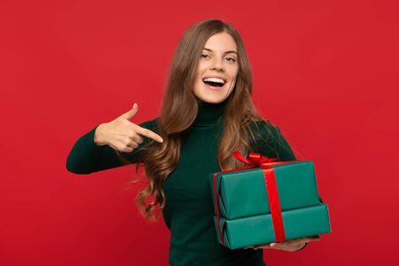 smiling woman in green sweater pointing on gift isolated on red background. Christmas conceptの写真素材