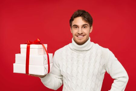 smiling man in white sweater holding Christmas presents isolated on red background. Christmas conceptの写真素材