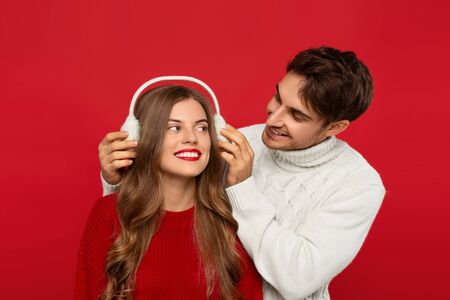 smiling man putting fur earmuffs on girlfriend head isolated on red background. Christmas conceptの写真素材