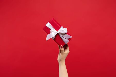 cropped view of woman holding wrapped Christmas present with white ribbon in hand isolated on red background. Christmas conceptの写真素材