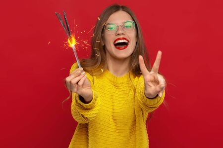 selective focus of happy girl in eyeglasses holding sparklers and showing peace sign isolated on redの写真素材