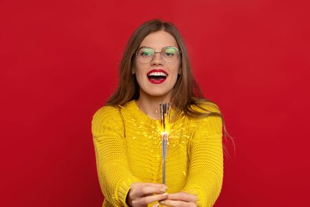 excited girl in eyeglasses holding sparklers, wears yellow sweater, looks with happiness, expresses good emotions, isolated on red background. Christmas conceptの写真素材