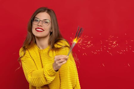 happy girl in eyeglasses holding sparklers, wears knitted yellow jumper, smiles pleasantly, has natural beauty, isolated on red background. People, clothes, warmthの写真素材