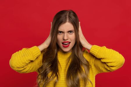 angry pretty girl in yellow sweater with red lips covering ears with hands isolated on red background. Face expressions conceptの写真素材