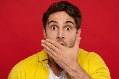 shocked man in yellow shirt covering mouth, looks surprisingly at camera isolated on red background. Horizontal shot of fascinated scared guy hears rumorの写真素材