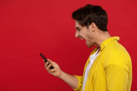 side view of angry man in yellow shirt screaming at smartphone, yells loudly, keeps mouth wide opened, frowns face in dissatisfaction isolated on red background. People, negative emotions and feelingsの写真素材