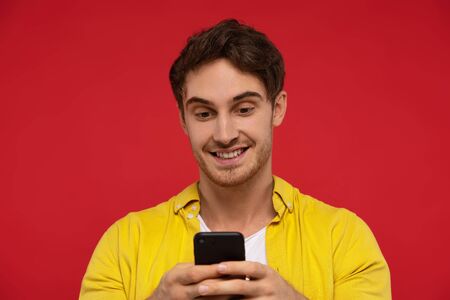 Portrait of happy man in yellow shirt using smartphone, smiles joyfully, isolated on red background. People and pastime conceptの写真素材
