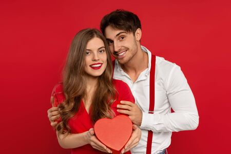 Cheerful nice young beautiful woman and man embrace in elegant outfit holding heart shaped box isolated on redの写真素材