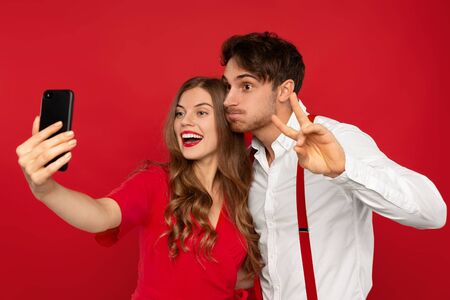 Smiling beautiful young couple in elegant outfits taking selfie and showing peace sign isolated on redの写真素材