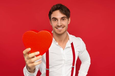 smiling handsome man in suspender and shirt holding heart shaped box isolated on red background, valentines conceptの写真素材