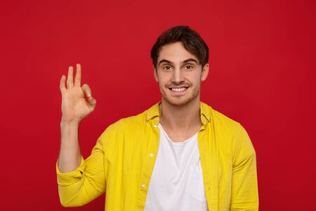 Yeah, its ok. Positive handsome man in yellow shirt showing ok sign with one hand, has big smile, being in high spirit, isolated on red backgroundの写真素材