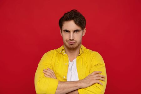 offended handsome man in yellow shirt with crossed arms isolated on red backgroundの写真素材