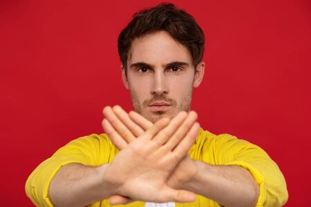 serious man in yellow shirt showing stop gesture at camera isolated on red backgroundの写真素材
