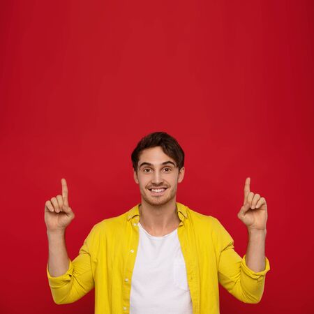 Cheerful handsome man in yellow shirt with amazed happy expression pointing with fingers up isolated on red background. Man in pleased to notice somethingの写真素材