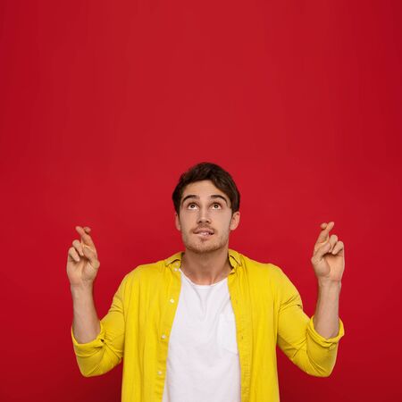 handsome man in yellow shirt with crossed fingers looking up, needs fortune during passing examination session isolated on red backgroundの写真素材