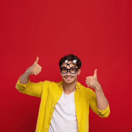 happy funny man in yellow shirt with many eyeglasses on face showing thumbs up isolated on red backgroundの写真素材