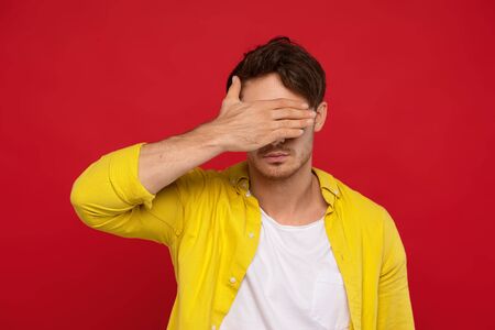 handsome man in yellow shirt covering face with hand isolated on red backgroundの写真素材