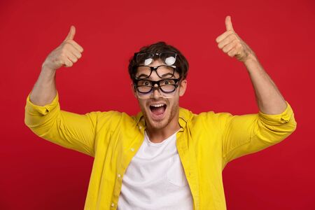 happy funny man in yellow shirt with many eyeglasses on face showing thumbs up isolated on red backgroundの写真素材