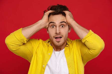 crazy man in yellow shirt holding head and looking at camera isolated on red backgroundの写真素材