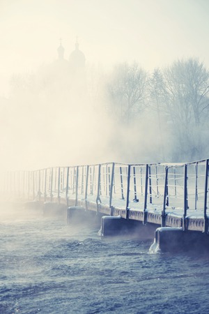 old bridge over the river in a frosty morning with the evaporation of water  background - silhouette of the domes of the churchの写真素材