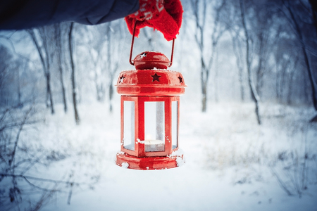 Holding in red glove a red candle lantern in the winter forest. Snow forest snowfall. Christmas Winter New Year background trembling flame Scenery. Fairy magic forest flashlightの写真素材