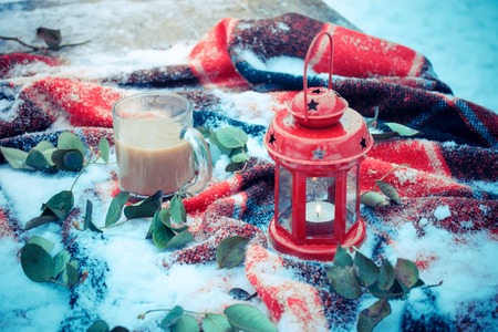 Festive red candle in a lantern and a mug of coffee on a red rug with snow with green leaves. Winter snow Christmas New Year holiday background snowy winter filterの写真素材