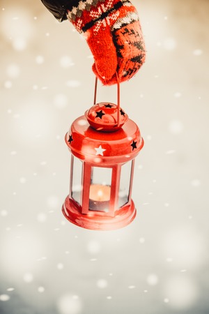 The girl in the red gloves holding a red lantern candle on a background of white snow in winter during a snowfallの写真素材