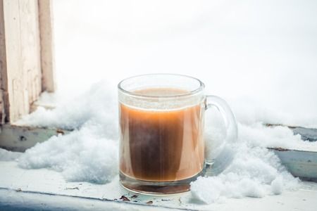 hot coffee with milk in a glass mug, which is studded with snow on the windowsill on window in winter cloudy day close-upの写真素材