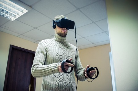 Man in helmet virtual reality plays game. Man uses VR-headset display with headphones for virtual reality game in office. High-tech devices. Augmented reality device creating virtual spaceの写真素材