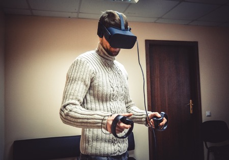 Man in helmet virtual reality plays game. Man uses VR-headset display with headphones for virtual reality game in office. High-tech devices. Augmented reality device creating virtual spaceの写真素材