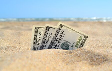Money dolars half covered with sand lie on sandy beach near sea ocean waves on sunny summer day close-up. Money grows out of the ground. Concept finance money holiday relax vacation.の写真素材